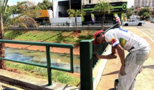 Trabalhador de boné vermelho pinta de verde o guarda-corpo de uma ponte sobre o canal da Avenida Aurora, em Olímpia