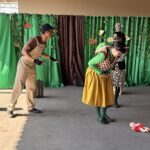 Atores de uma peça de teatro sobre bullying em cena diante de cortinas verdes, com uma boneca caída no chão