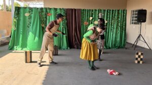 Atores de uma peça de teatro sobre bullying em cena diante de cortinas verdes, com uma boneca caída no chão