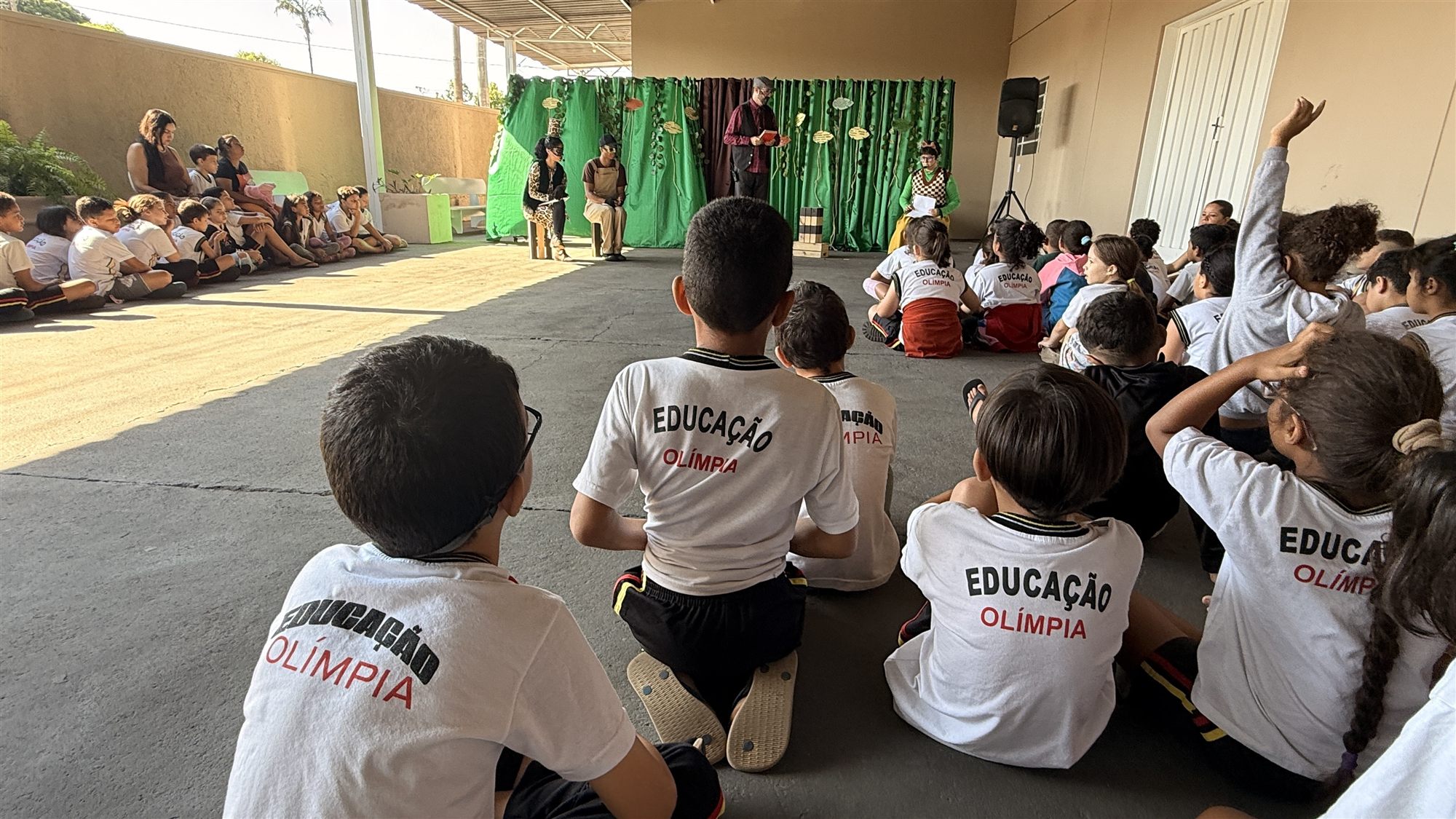 Crianças de camisetas brancas do uniforme escolar sentadas no chão assistem a uma peça de teatro, com uma menina de braço levantado