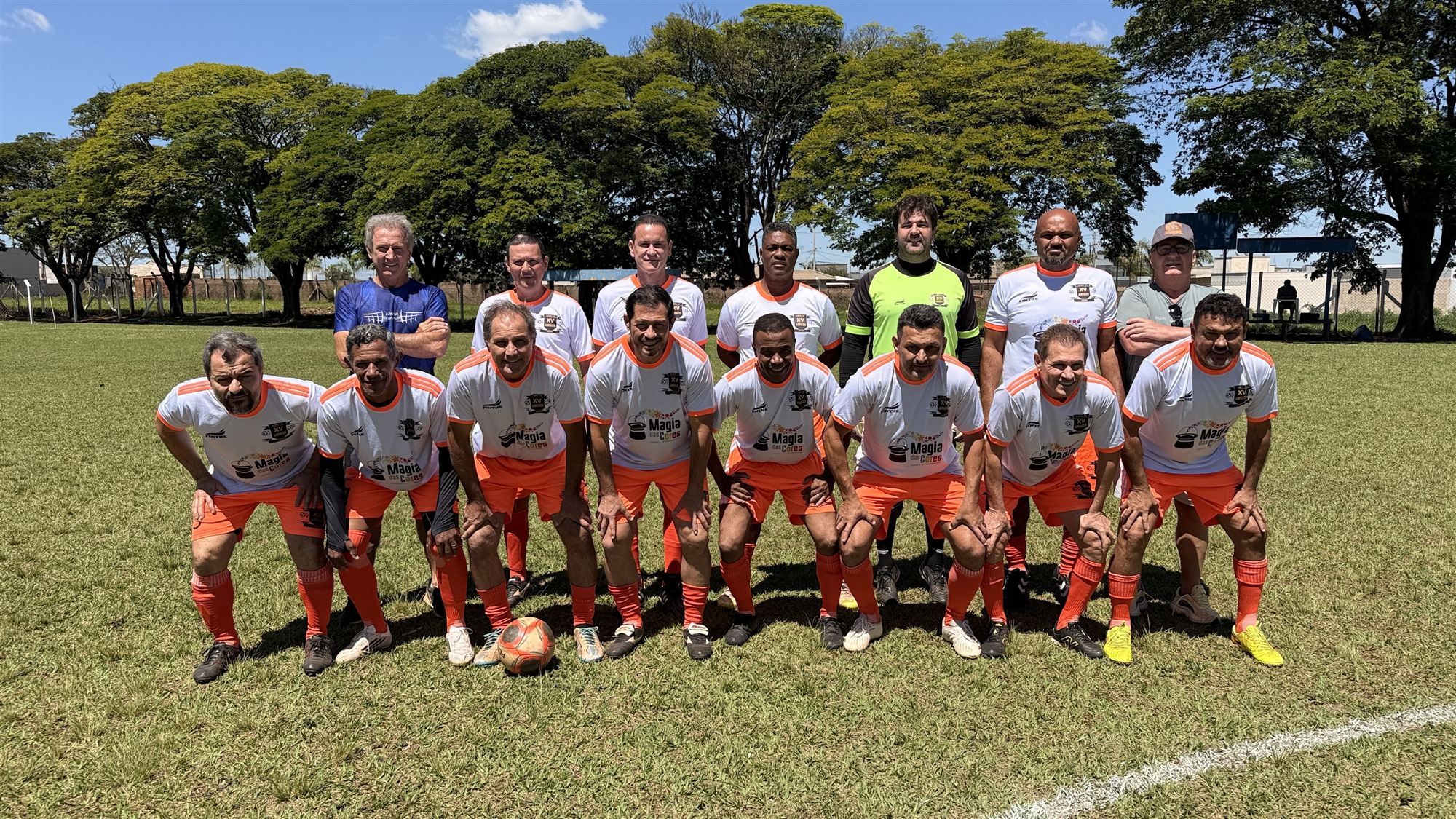 Equipe de camisas brancas com detalhes laranja posa para foto com a bola no campo do Campeonato Veterano de Futebol de Olímpia