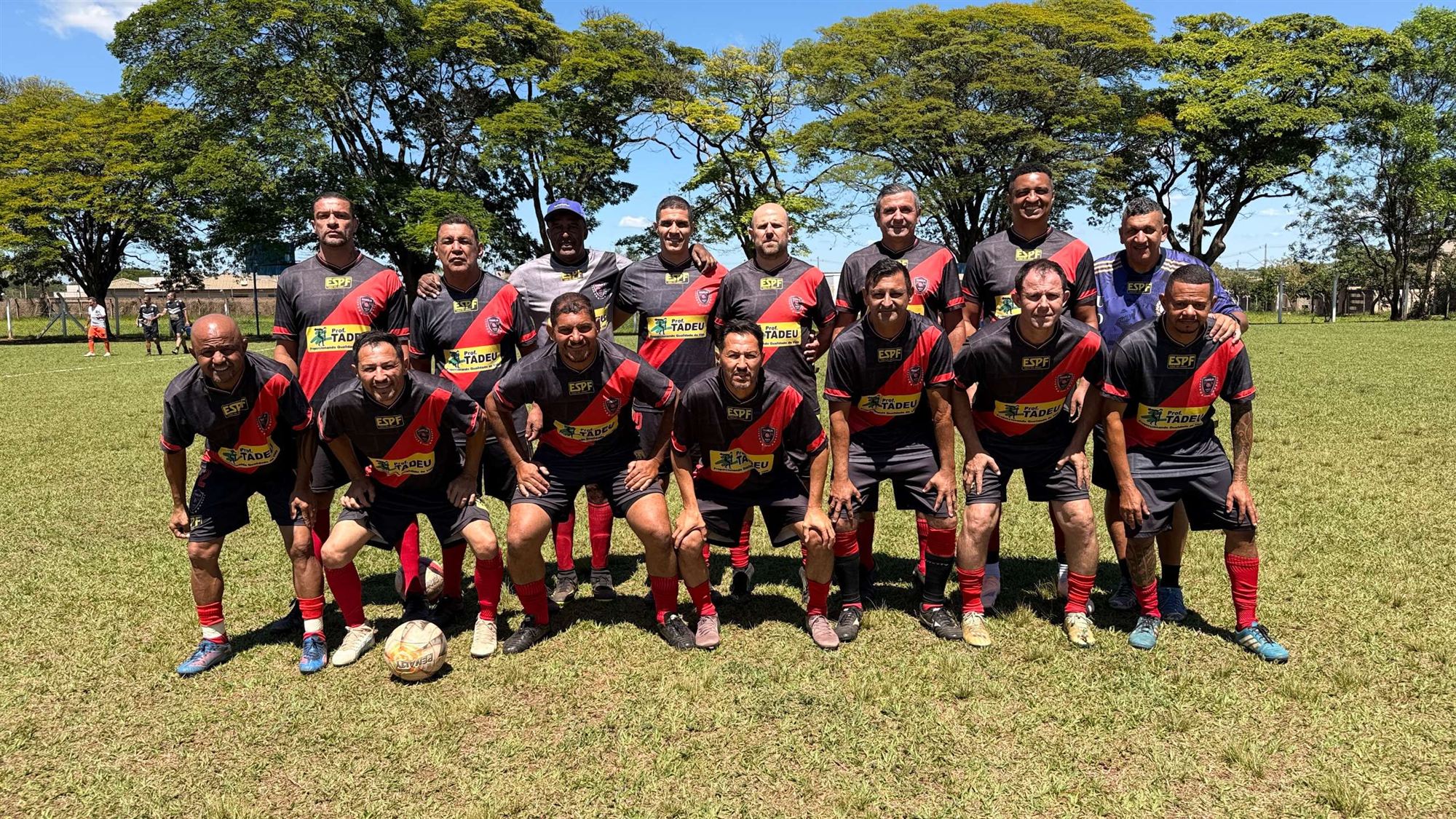 Equipe de camisas pretas com faixa diagonal vermelha posa para foto no campo durante o Campeonato Veterano de Futebol de Olímpia