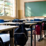 Sala de aula de escola pública vazia, com carteiras enfileiradas, mochilas penduradas nas cadeiras e lousa verde limpa