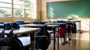 Sala de aula de escola pública vazia, com carteiras enfileiradas, mochilas penduradas nas cadeiras e lousa verde limpa