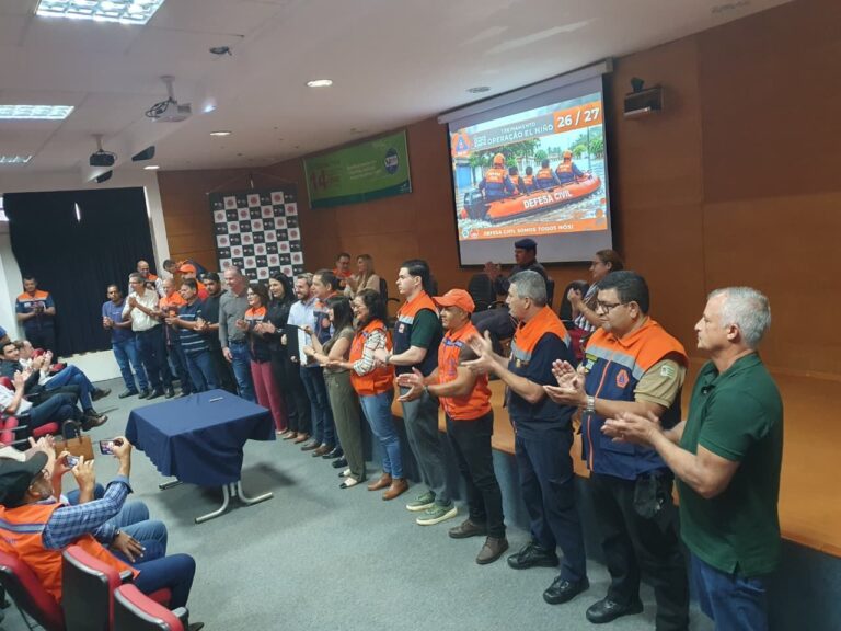 Participantes de treinamento da Defesa Civil em um auditório, em pé e aplaudindo, com telão da Operação El Niño ao fundo