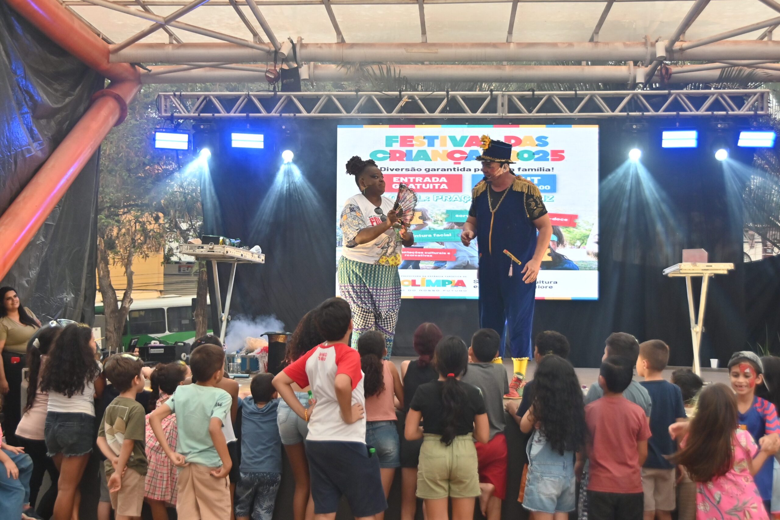 Dois artistas se apresentam em um palco diante de crianças que assistem de pé, em evento infantil em Olímpia