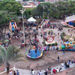 Vista aérea de praça de Olímpia com brinquedos infláveis, cama elástica e famílias reunidas em evento para crianças