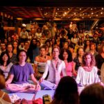 Plateia sentada de olhos fechados em meditação coletiva ao som de tambores, em salão com iluminação quente