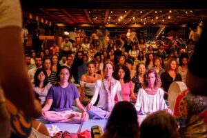 Plateia sentada de olhos fechados em meditação coletiva ao som de tambores, em salão com iluminação quente