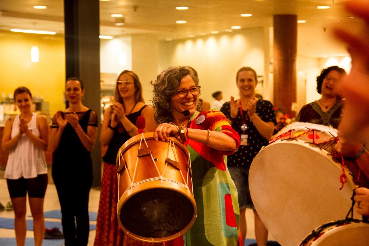 Mulher sorri e segura um tambor de madeira durante atividade de meditação com tambores, aplaudida por participantes