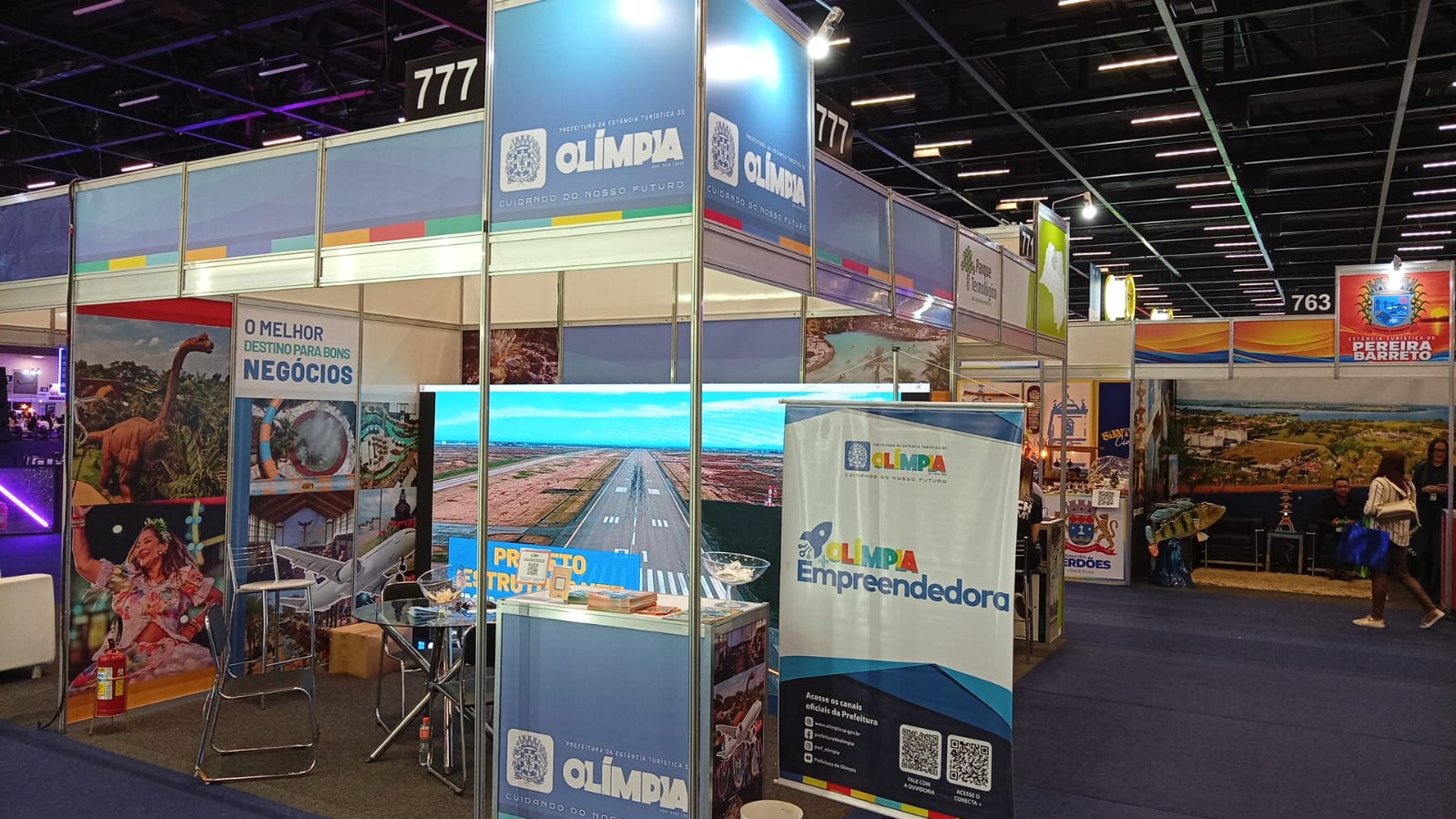 Estande de Olímpia na Feira do Empreendedor 2026, em São Paulo, com painéis da cidade, telão com imagem do aeroporto e banner do programa Olímpia Empreendedora