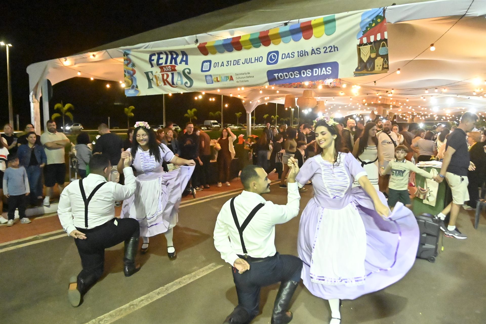 Dançarinos de traje a caráter se apresentam sob a tenda da Feira nas Férias, com público ao redor, em Olímpia