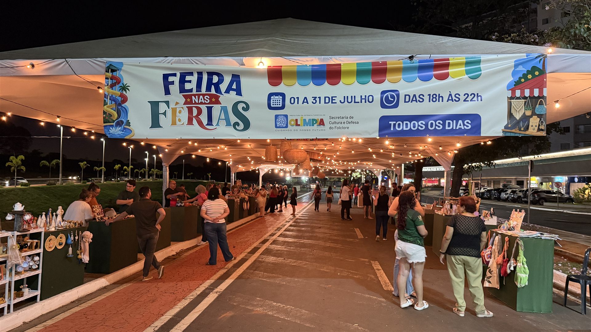 Tenda iluminada com barracas de artesanato e público circulando na Feira nas Férias, na avenida turística de Olímpia