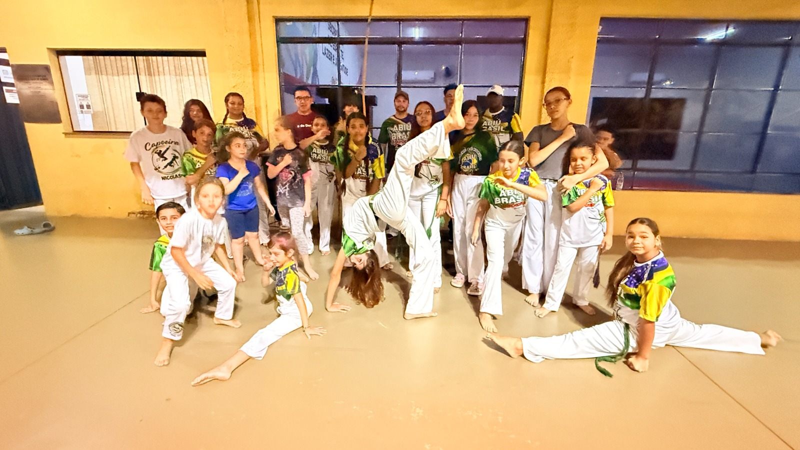 Grupo de crianças e adultos de uniforme branco posa em sala depois da apresentação de capoeira