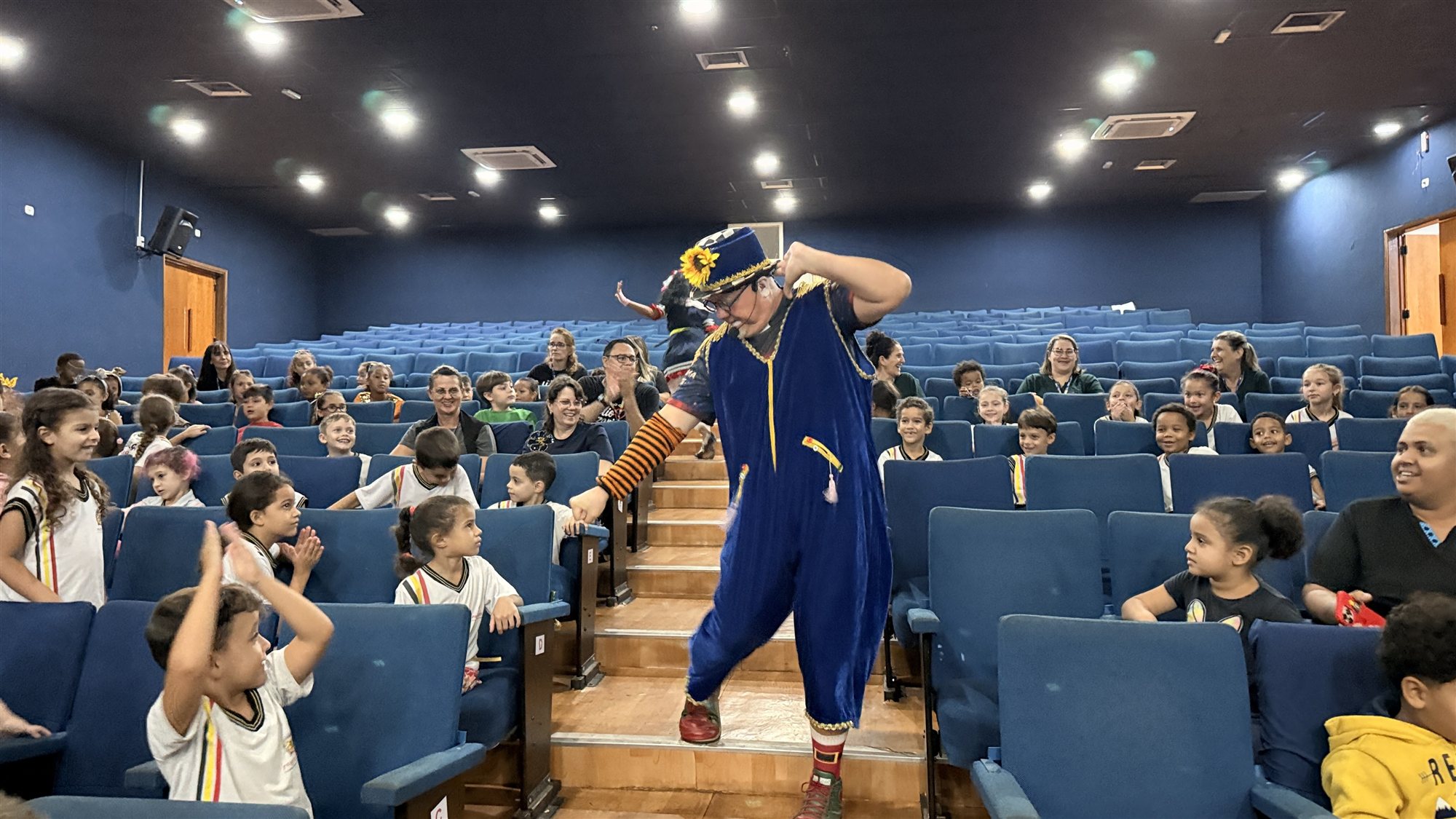 Palhaço de macacão azul desce a escada de um teatro entre crianças de uniforme que sorriem e batem palmas