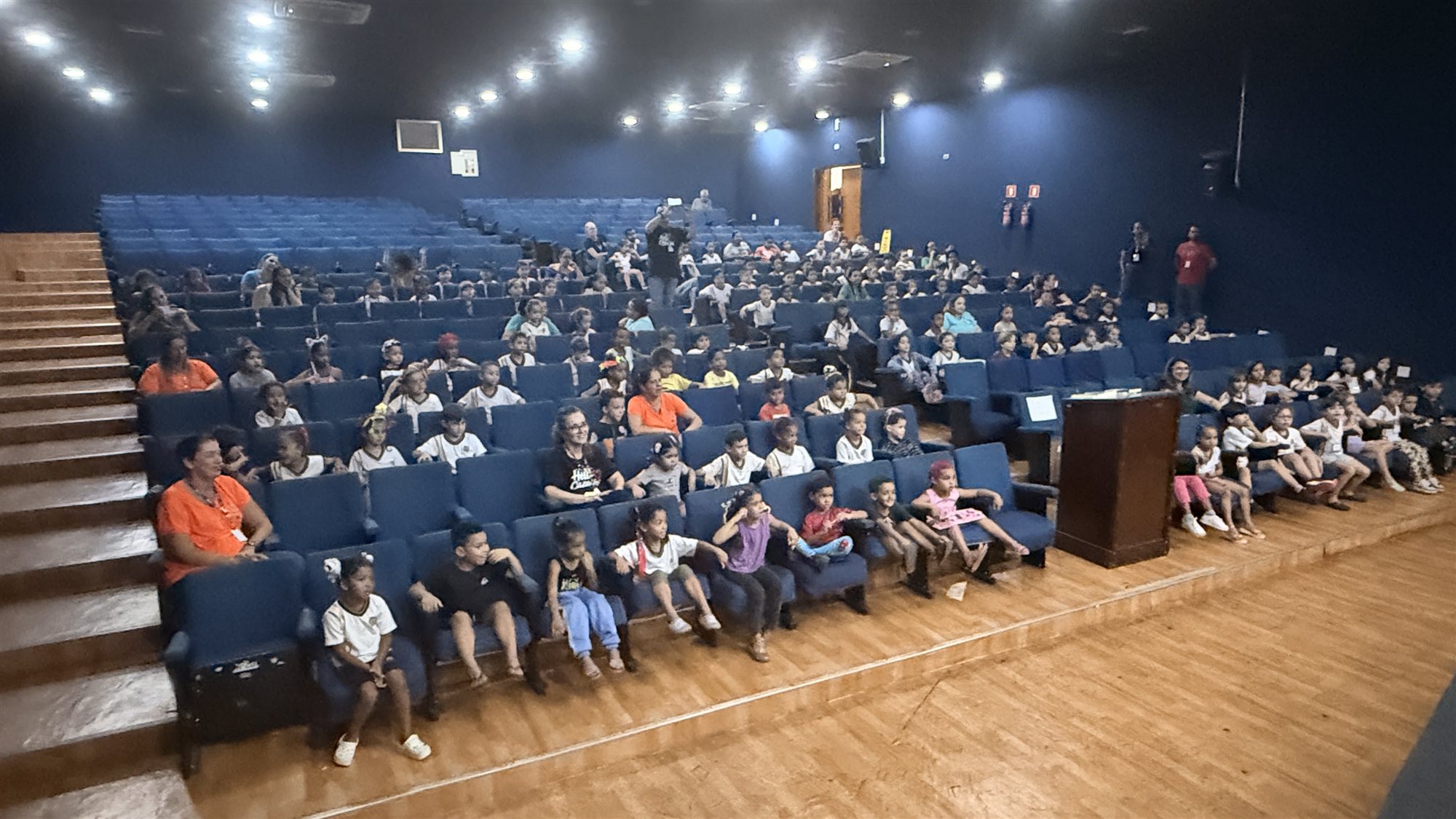 Plateia de crianças de uniforme e professoras sentadas nas poltronas azuis da Casa da Cultura de Olímpia