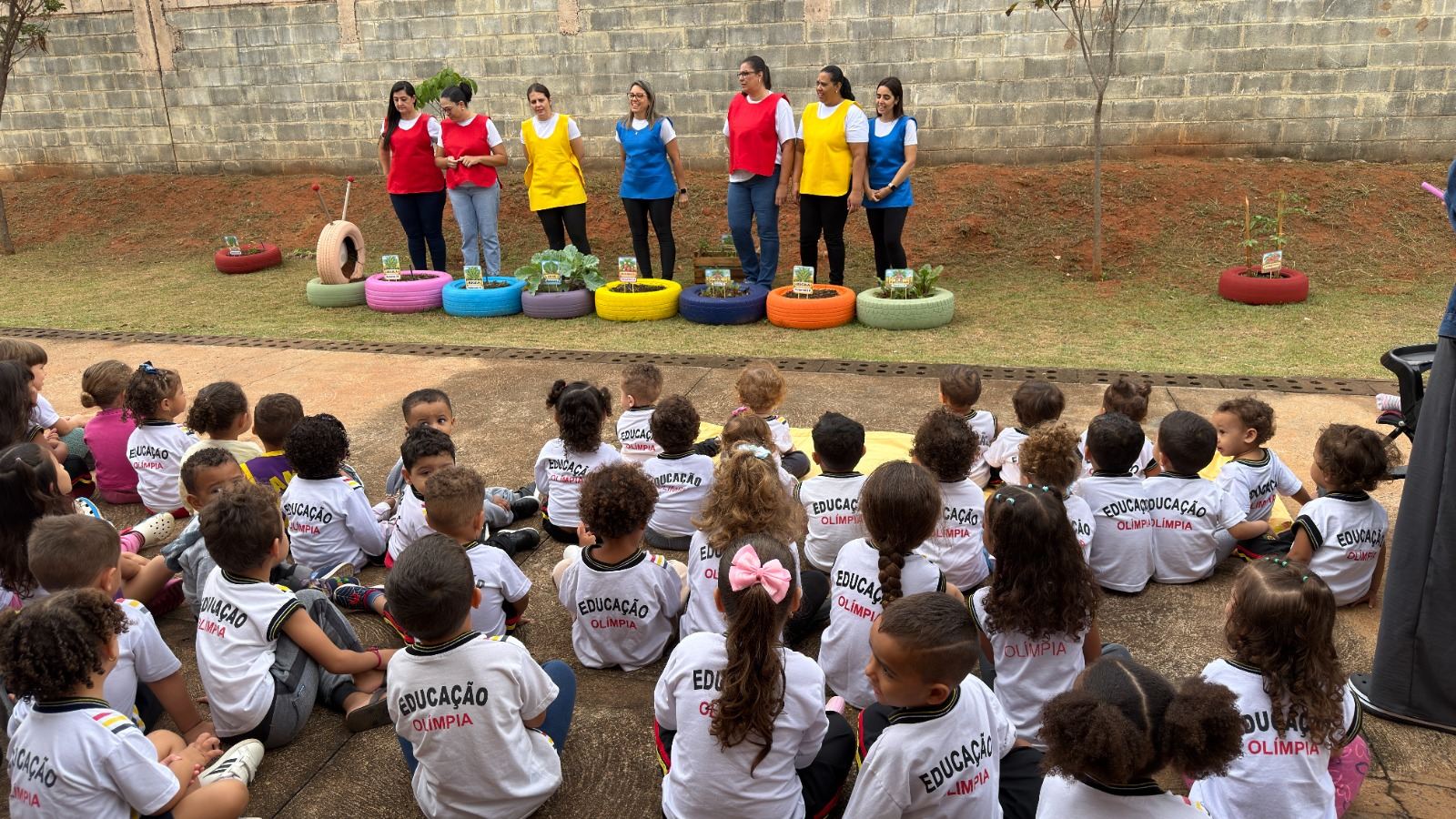 Crianças uniformizadas sentadas no chão, de frente para profissionais da escola e para canteiros feitos de pneus coloridos