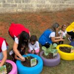 Professoras e crianças plantam mudas em pneus pintados de cores diferentes, usados como canteiros na horta da escola