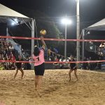 Jogadoras disputam partida de vôlei de areia à noite, com o público nas arquibancadas ao fundo