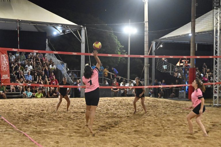 Jogadoras disputam partida de vôlei de areia à noite, com o público nas arquibancadas ao fundo