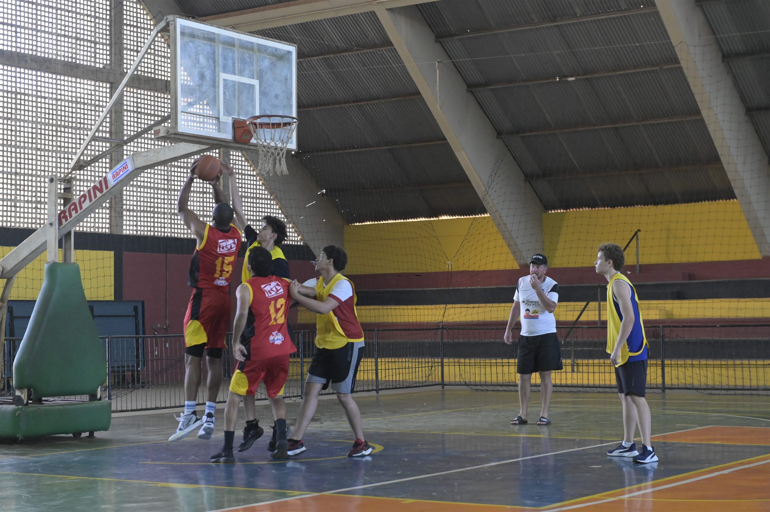 Jogadores disputam uma partida de basquete debaixo da cesta em quadra coberta, com um árbitro acompanhando o lance