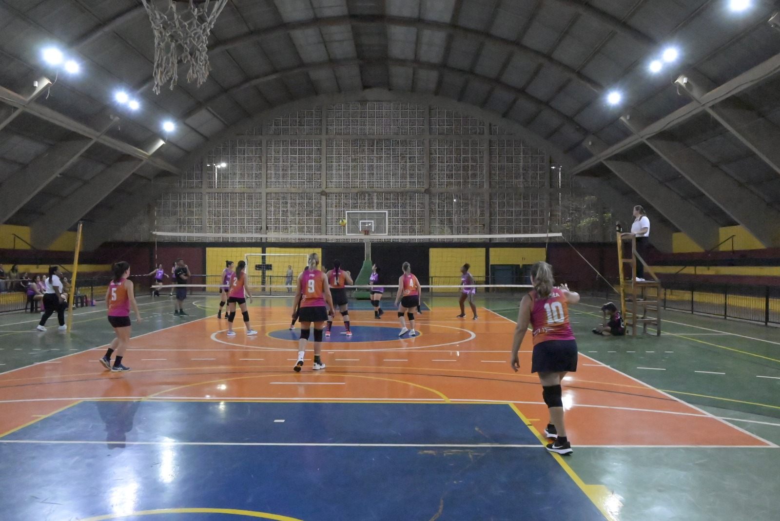 Mulheres de camisetas rosa e laranja jogam vôlei em quadra coberta, com a rede ao fundo e uma árbitra na cadeira de arbitragem