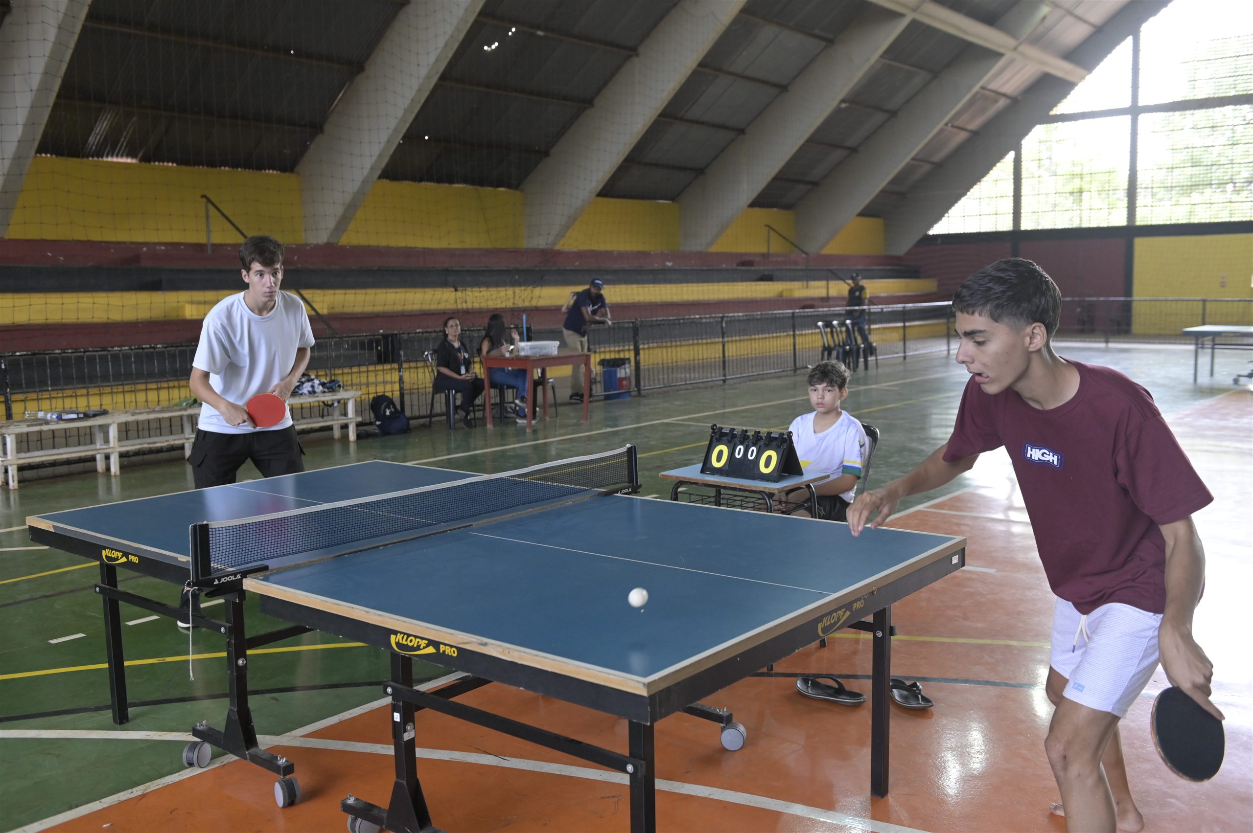 Dois jogadores disputam uma partida de tênis de mesa em ginásio, com um menino sentado ao fundo marcando o placar