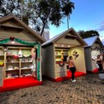 Casinhas de madeira decoradas com luzes e enfeites de Natal, com visitantes olhando os produtos, na Praça da Matriz, em Olímpia
