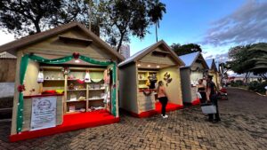 Casinhas de madeira decoradas com luzes e enfeites de Natal, com visitantes olhando os produtos, na Praça da Matriz, em Olímpia