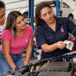 Instrutora mostra a vareta de óleo a duas mulheres diante do motor aberto de um carro, em uma oficina; as três usam o laço rosa do Outubro Rosa