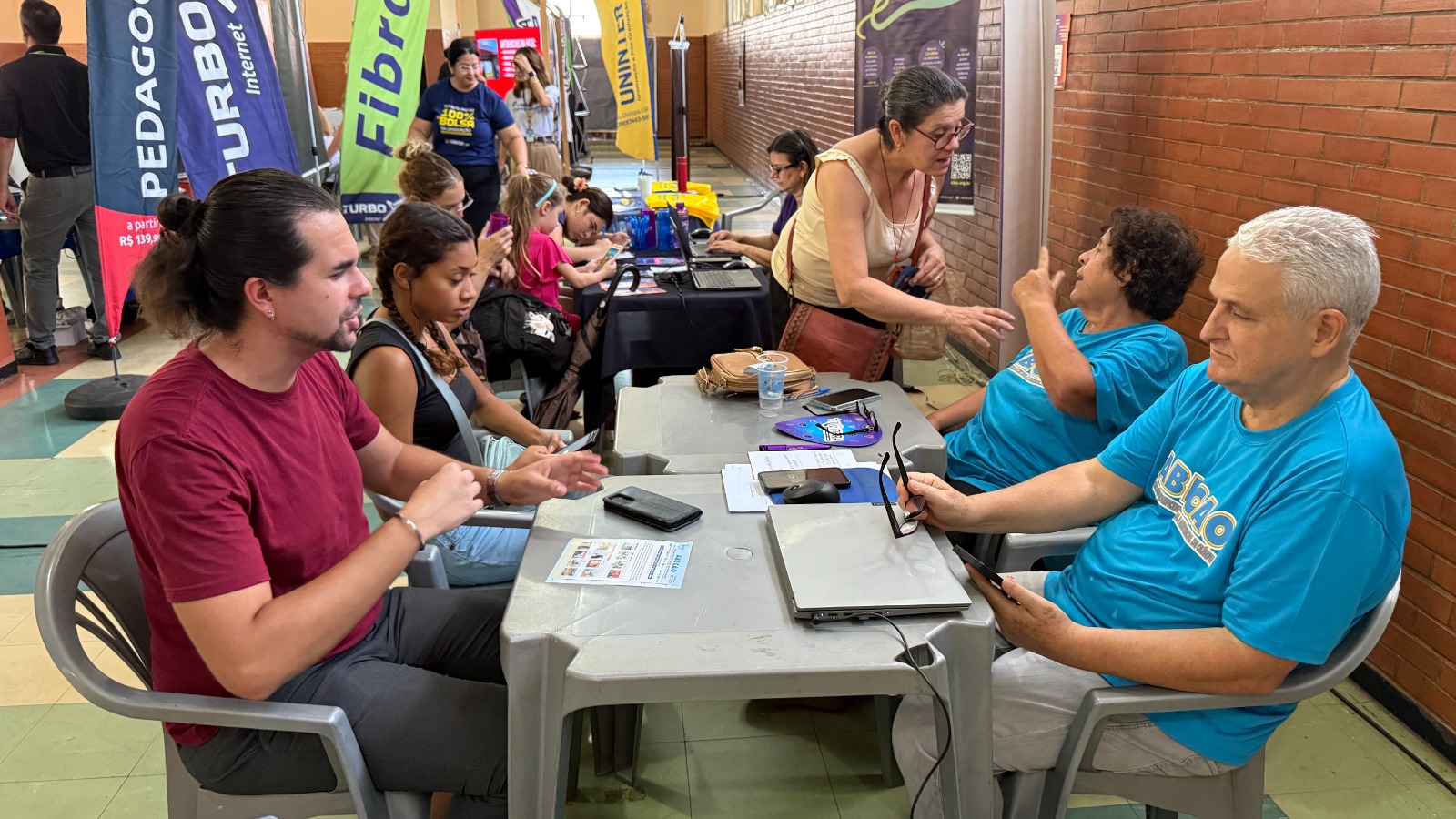 Candidatos e atendentes de entidade conversam em mesas, com faixas de empresas e instituições de ensino ao fundo, em mutirão de emprego