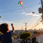Criança de costas empina pipa colorida em rua de bairro, com fios e poste de energia à direita e pôr do sol ao fundo