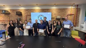 Grupo reunido em sala com tijolos aparentes, segurando certificados do selo Empresa Amiga do Autista, com o logotipo do programa ao fundo