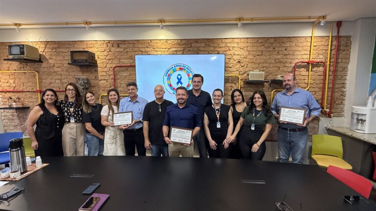 Grupo reunido em sala com tijolos aparentes, segurando certificados do selo Empresa Amiga do Autista, com o logotipo do programa ao fundo