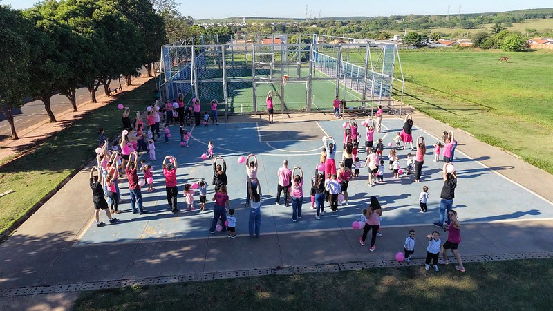Vista aérea de crianças e adultos de rosa fazendo ginástica em uma quadra, com balões