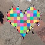 Vista aérea de um grande coração de tecido colorido aberto no chão de uma praça, cercado por pessoas de rosa