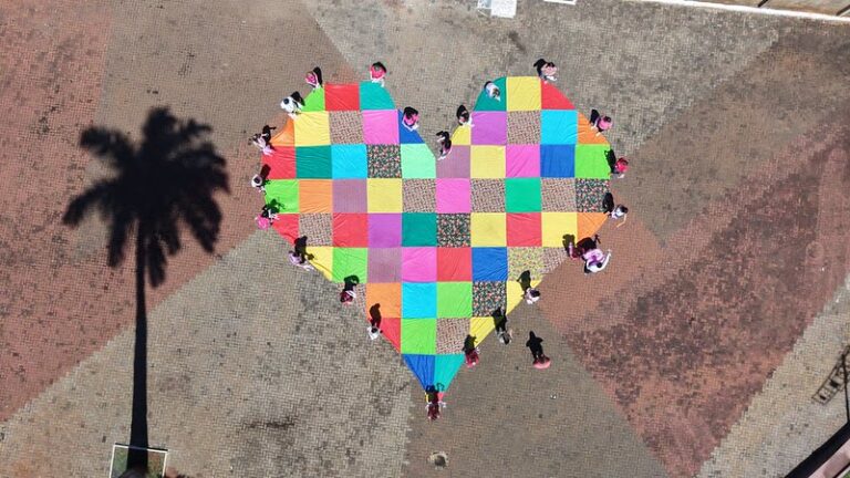 Vista aérea de um grande coração de tecido colorido aberto no chão de uma praça, cercado por pessoas de rosa
