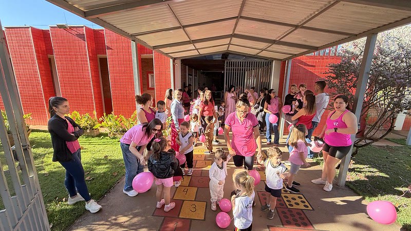 Crianças e funcionárias de rosa com balões no pátio de uma escola municipal