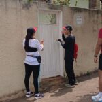 Três profissionais de saúde com bonés e mochilas vermelhas conversam com morador diante do portão de uma casa, em bairro de Olímpia