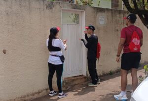 Três profissionais de saúde com bonés e mochilas vermelhas conversam com morador diante do portão de uma casa, em bairro de Olímpia