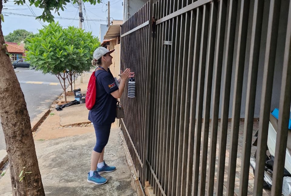 Agente de saúde de boné e mochila vermelha conversa com morador diante do portão de uma casa, durante a busca ativa por tuberculose em Olímpia